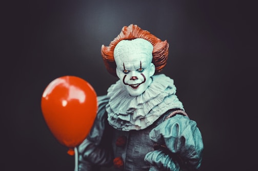A creepy clown with a white face, red nose, and exaggerated makeup holds a bright red balloon. The clown has orange hair and wears a ruffled gray outfit, set against a dark background.