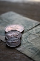 Three coins are placed on top of a paper currency with intricate designs. The focus is on the coins, which appear to have an embossed profile of a person and include engraved text.