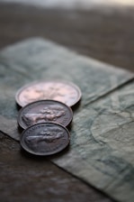 Three coins are placed on top of a paper currency with intricate designs. The focus is on the coins, which appear to have an embossed profile of a person and include engraved text.