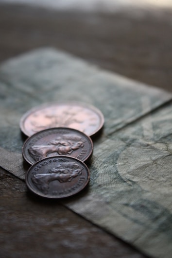 Three coins are placed on top of a paper currency with intricate designs. The focus is on the coins, which appear to have an embossed profile of a person and include engraved text.
