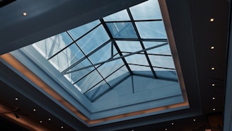 A modern architectural ceiling with a large, geometric glass skylight allowing natural light to illuminate the space. The ceiling has recessed lighting around its edges, giving a warm glow against the smooth surfaces.