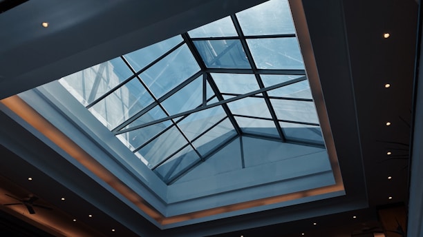 A modern architectural ceiling with a large, geometric glass skylight allowing natural light to illuminate the space. The ceiling has recessed lighting around its edges, giving a warm glow against the smooth surfaces.