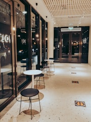 A modern hallway with large glass windows on the left and a row of small round tables and black chairs along the corridor. The ceiling features a grid design with recessed lighting, and the floor has a speckled pattern with signs indicating social distancing. Warm lighting creates a cozy atmosphere.