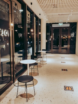 A modern hallway with large glass windows on the left and a row of small round tables and black chairs along the corridor. The ceiling features a grid design with recessed lighting, and the floor has a speckled pattern with signs indicating social distancing. Warm lighting creates a cozy atmosphere.