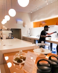 A barista wearing a black mask and gloves is preparing drinks in a clean and modern coffee shop. The counter is white and glossy with pendant lights hanging from the ceiling. There is a pastry display with cookies under glass domes and a menu stand on the counter. The background features orange and white colors.