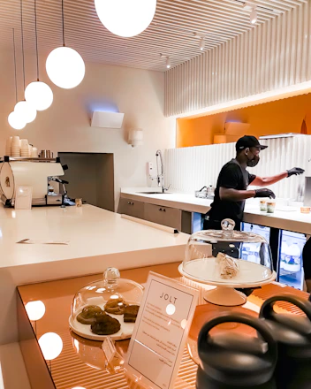 A barista wearing a black mask and gloves is preparing drinks in a clean and modern coffee shop. The counter is white and glossy with pendant lights hanging from the ceiling. There is a pastry display with cookies under glass domes and a menu stand on the counter. The background features orange and white colors.
