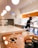 A barista wearing a black mask and gloves is preparing drinks in a clean and modern coffee shop. The counter is white and glossy with pendant lights hanging from the ceiling. There is a pastry display with cookies under glass domes and a menu stand on the counter. The background features orange and white colors.