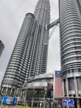 Two towering modern skyscrapers dominate the skyline, featuring a unique architectural design with cylindrical and reflective surfaces. The buildings are connected by a skybridge at higher levels. Large Malaysian flags are prominently displayed, and the surroundings include palm trees and urban signage.