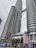 Two towering modern skyscrapers dominate the skyline, featuring a unique architectural design with cylindrical and reflective surfaces. The buildings are connected by a skybridge at higher levels. Large Malaysian flags are prominently displayed, and the surroundings include palm trees and urban signage.
