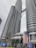 Two towering modern skyscrapers dominate the skyline, featuring a unique architectural design with cylindrical and reflective surfaces. The buildings are connected by a skybridge at higher levels. Large Malaysian flags are prominently displayed, and the surroundings include palm trees and urban signage.