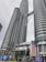 Two towering modern skyscrapers dominate the skyline, featuring a unique architectural design with cylindrical and reflective surfaces. The buildings are connected by a skybridge at higher levels. Large Malaysian flags are prominently displayed, and the surroundings include palm trees and urban signage.