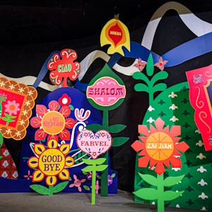 A colorful display of stylized flowers, each featuring different words for 'goodbye' in various languages, including 'Ciao,' 'Shalom,' 'Au Revoir,' 'Good Bye,' 'Adeus,' 'Farvel,' and 'Zai Jian.' The flowers are highly decorative, surrounded by vibrant colors and patterns, and are set against a dark background with an abstract mountain and wave-like elements.
