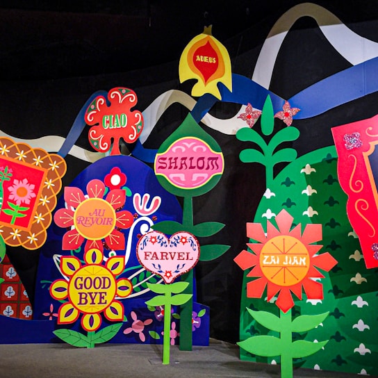 A colorful display of stylized flowers, each featuring different words for 'goodbye' in various languages, including 'Ciao,' 'Shalom,' 'Au Revoir,' 'Good Bye,' 'Adeus,' 'Farvel,' and 'Zai Jian.' The flowers are highly decorative, surrounded by vibrant colors and patterns, and are set against a dark background with an abstract mountain and wave-like elements.