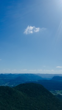 A single cloud floats in a clear blue sky above a scenic landscape with rolling, forested hills that extend into the distance. The sunlight creates a serene and bright atmosphere.