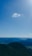 A single cloud floats in a clear blue sky above a scenic landscape with rolling, forested hills that extend into the distance. The sunlight creates a serene and bright atmosphere.