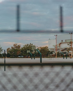 A basketball court is visible through a chain-link fence with several basketball hoops and backboards. The area is surrounded by trees and a large building is seen in the background. The sky has a mix of clouds, creating a serene atmosphere.