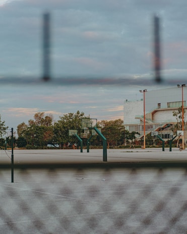 A basketball court is visible through a chain-link fence with several basketball hoops and backboards. The area is surrounded by trees and a large building is seen in the background. The sky has a mix of clouds, creating a serene atmosphere.