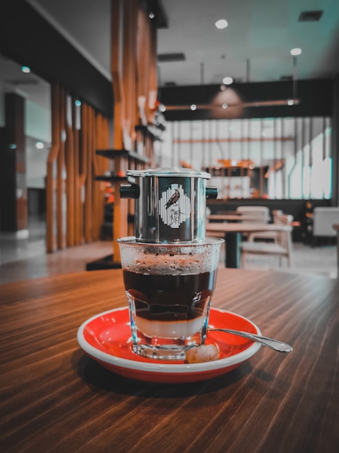 A glass of coffee with milk served on a red saucer sits on a wooden table. A stainless steel drip filter is placed on top of the glass. The background consists of a modern, softly lit caf&eacute; with wooden and metal elements, including tables and chairs.