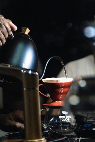 A person is pouring hot water from a black kettle into a red pour-over coffee dripper, which is positioned on top of a glass carafe labeled 'one two cups'. The scene is set in a dark, possibly indoor environment with focused lighting on the brewing process.