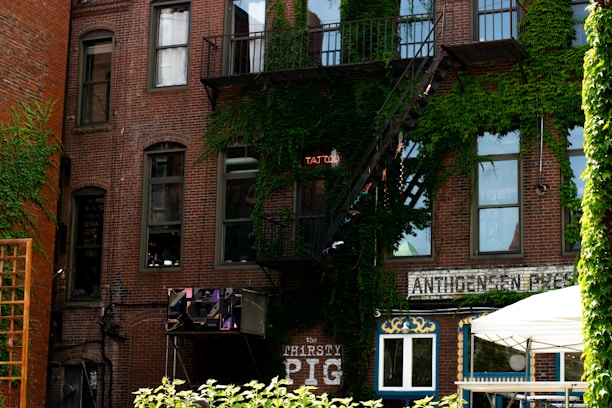 A brick building with multiple windows, partly covered by green ivy. A metal fire escape leads to the upper stories. A neon sign reads 'TATTOO', and a business sign for 'The THIRSTY PIG' is visible. A white canopy tent is set up in front of the building.