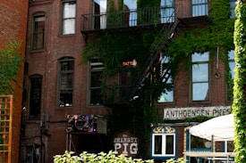 A brick building with multiple windows, partly covered by green ivy. A metal fire escape leads to the upper stories. A neon sign reads 'TATTOO', and a business sign for 'The THIRSTY PIG' is visible. A white canopy tent is set up in front of the building.