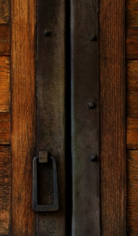 A close-up view of a rustic wooden door featuring a large strip of dark, weathered metal running vertically down its center. Affixed to the metal strip is a rectangular, heavy iron handle designed for pulling. The wood displays a rich, natural grain with tones of brown and hints of aged distress.