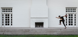 A person is captured mid-air while performing a dynamic leap off a set of steps, in front of a symmetrical building facade with two doors and a central fireplace. The scene exudes a sense of motion and energy against the minimalist backdrop.