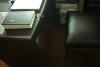 A dark wooden desk with a closed black book placed on top, featuring gold lettering and a red ribbon bookmark hanging out. Nearby is a closed laptop, partially visible, and a dark leather chair at the side. In the background, there are shelves with various papers and books.