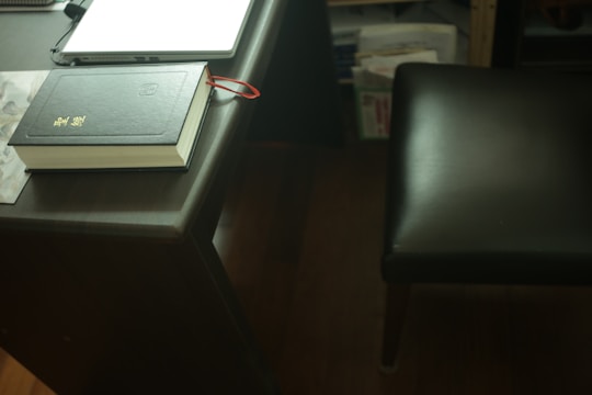 A dark wooden desk with a closed black book placed on top, featuring gold lettering and a red ribbon bookmark hanging out. Nearby is a closed laptop, partially visible, and a dark leather chair at the side. In the background, there are shelves with various papers and books.