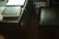 A dark wooden desk with a closed black book placed on top, featuring gold lettering and a red ribbon bookmark hanging out. Nearby is a closed laptop, partially visible, and a dark leather chair at the side. In the background, there are shelves with various papers and books.