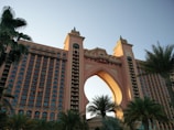 A large, luxurious hotel with a grand arch at its center, flanked by two towers featuring numerous windows. The building is surrounded by lush palm trees and bathed in soft sunlight, suggesting a location likely in a coastal or tropical region.