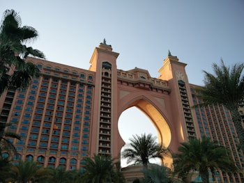 A large, luxurious hotel with a grand arch at its center, flanked by two towers featuring numerous windows. The building is surrounded by lush palm trees and bathed in soft sunlight, suggesting a location likely in a coastal or tropical region.