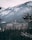 A large, historic hotel with a Canadian flag is nestled among snow-covered trees and mountainous terrain. The hotel features a classic architectural style with numerous windows and gabled roofs, set against a backdrop of rugged, snow-dusted mountains.