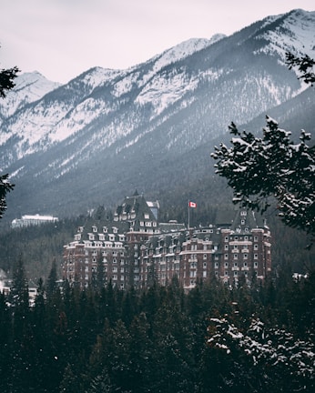 A large, historic hotel with a Canadian flag is nestled among snow-covered trees and mountainous terrain. The hotel features a classic architectural style with numerous windows and gabled roofs, set against a backdrop of rugged, snow-dusted mountains.