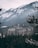 A large, historic hotel with a Canadian flag is nestled among snow-covered trees and mountainous terrain. The hotel features a classic architectural style with numerous windows and gabled roofs, set against a backdrop of rugged, snow-dusted mountains.