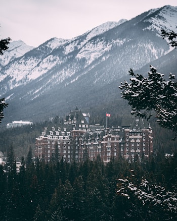 A large, historic hotel with a Canadian flag is nestled among snow-covered trees and mountainous terrain. The hotel features a classic architectural style with numerous windows and gabled roofs, set against a backdrop of rugged, snow-dusted mountains.