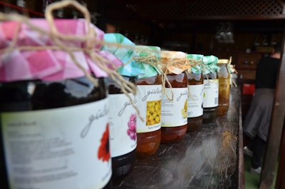 Jars of preserves with colorful fabric covers and twine ties are neatly arranged on a wooden shelf. Each jar has a label indicating the type of preserve inside, with fruits like lemons and other assortments depicted. The setting suggests a rustic, market-like environment.