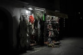 A dimly lit souvenir shop with various items such as clothing, fabrics, and traditional Croatian textiles hanging from racks. The shop exterior is illuminated by a single overhead light, casting shadows and highlighting the vibrant patterns and textures of the merchandise.