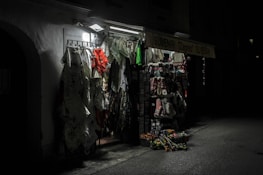 A dimly lit souvenir shop with various items such as clothing, fabrics, and traditional Croatian textiles hanging from racks. The shop exterior is illuminated by a single overhead light, casting shadows and highlighting the vibrant patterns and textures of the merchandise.