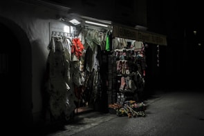 A dimly lit souvenir shop with various items such as clothing, fabrics, and traditional Croatian textiles hanging from racks. The shop exterior is illuminated by a single overhead light, casting shadows and highlighting the vibrant patterns and textures of the merchandise.