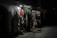 A dimly lit souvenir shop with various items such as clothing, fabrics, and traditional Croatian textiles hanging from racks. The shop exterior is illuminated by a single overhead light, casting shadows and highlighting the vibrant patterns and textures of the merchandise.