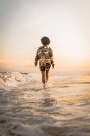 back of person in shorts and long-sleeve shirt walking at dusk through ankle-deep water at a beach 