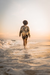 back of person in shorts and long-sleeve shirt walking at dusk through ankle-deep water at a beach 