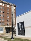 A historic hotel building with a brown brick facade and multiple windows. A black and white mural features a portrait of a man in a suit on an adjacent white wall. A street lamp is in the foreground, and a blue street sign is visible at a street corner. Scaffolding is attached to the hotel’s exterior, suggesting ongoing renovations. A blue sky provides a clear backdrop.