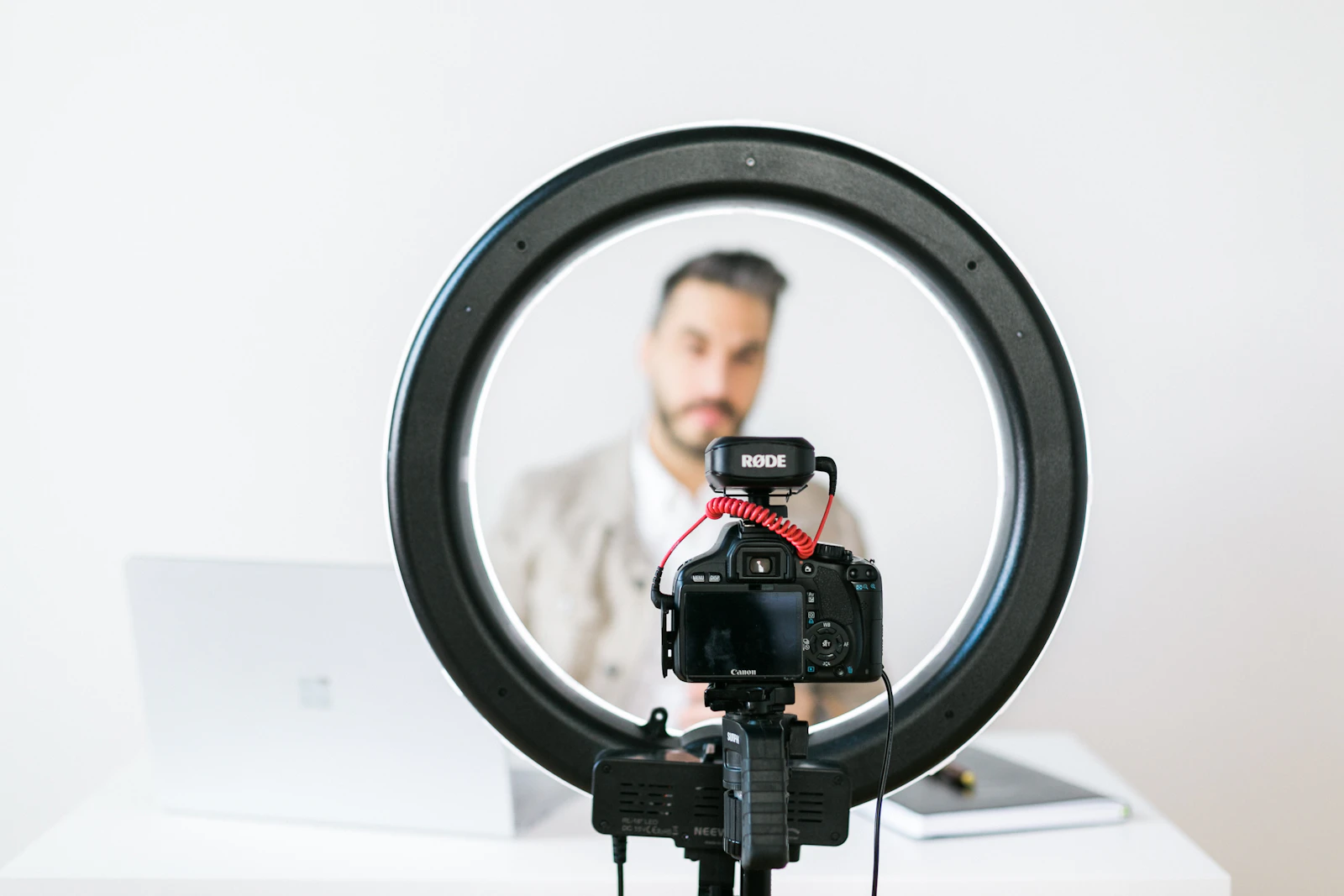 A camera mounted inside a ring light on a desk, pointed at a man in a white studio