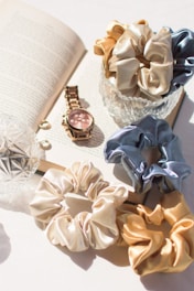 An open book serves as the backdrop for an arrangement of accessories. A gold watch with a pink face rests alongside the book, accompanied by two small round earrings. Several silk scrunchies in shades of gold, cream, and blue are artfully placed around a glass dish.