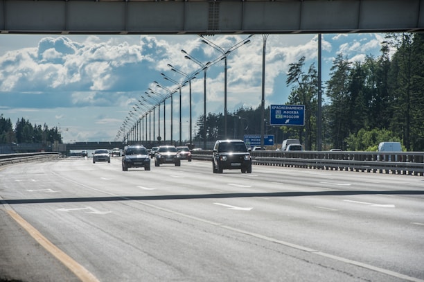 An open highway with multiple cars driving under an overpass. The sky is partly cloudy, and there are trees lining both sides of the road. A road sign is visible, providing directions to Moscow.