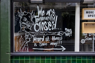 A storefront window features handwritten chalk-like artwork and text informing passersby that the business is temporarily closed and directing them to another location on Meard Street. The illustration includes the figure of a whimsical animal pointing to the right, accompanied by additional text to guide customers. There are elements of urban architecture visible, such as green tiles and part of a sign indicating directions to an upstairs area.