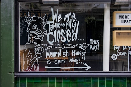 A storefront window features handwritten chalk-like artwork and text informing passersby that the business is temporarily closed and directing them to another location on Meard Street. The illustration includes the figure of a whimsical animal pointing to the right, accompanied by additional text to guide customers. There are elements of urban architecture visible, such as green tiles and part of a sign indicating directions to an upstairs area.