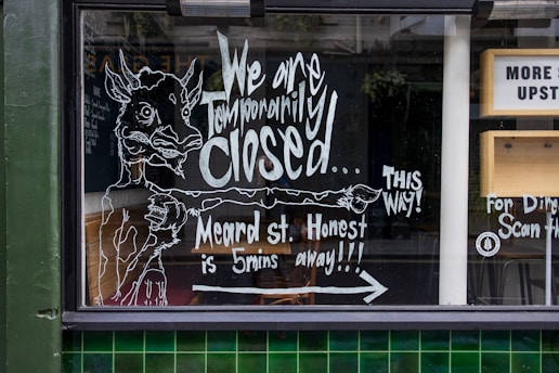 A storefront window features handwritten chalk-like artwork and text informing passersby that the business is temporarily closed and directing them to another location on Meard Street. The illustration includes the figure of a whimsical animal pointing to the right, accompanied by additional text to guide customers. There are elements of urban architecture visible, such as green tiles and part of a sign indicating directions to an upstairs area.